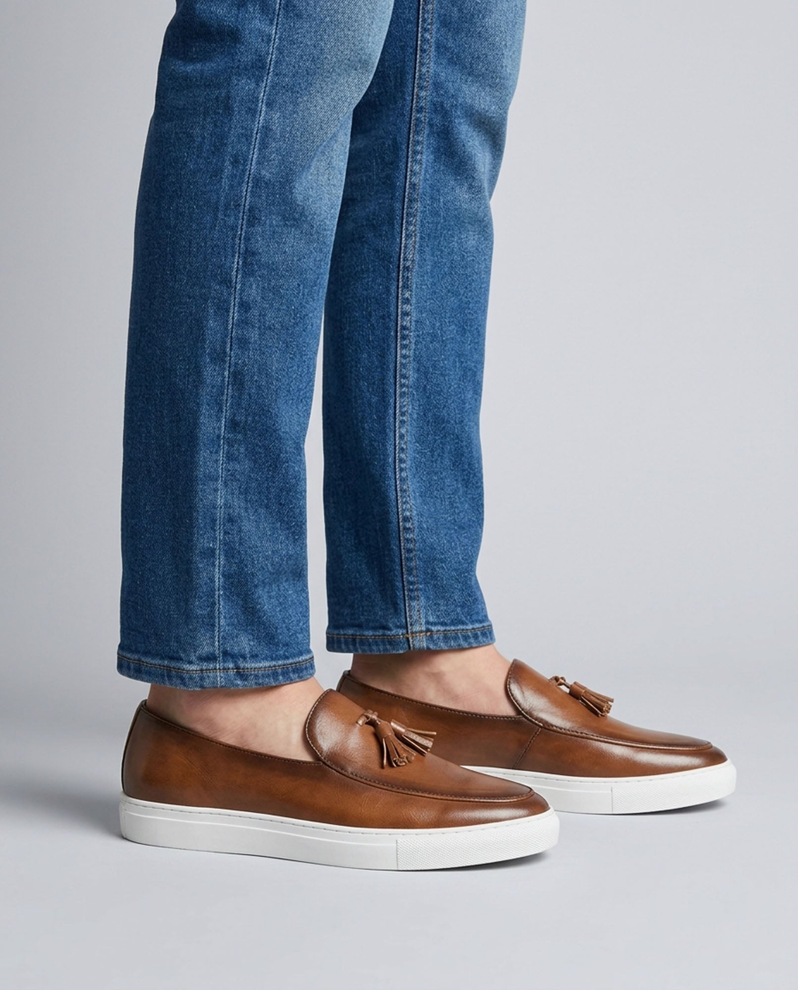 Capri men's cognac leather tassel loafers styled with blue denim jeans on grey background
