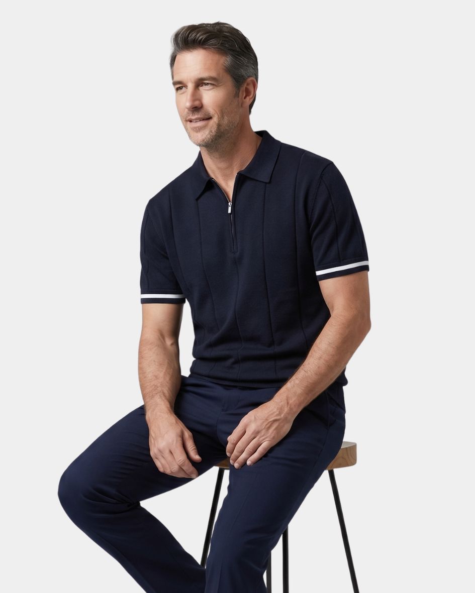 Man wearing a navy polo shirt and pants sitting on a stool against a white background