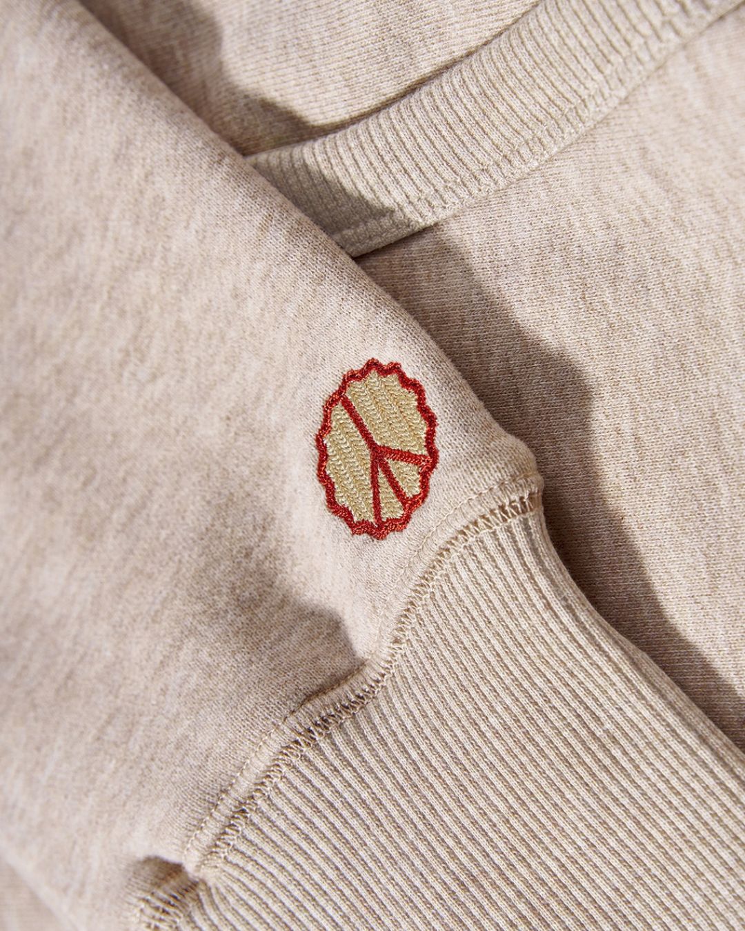 Beige cotton hoodie sleeve with a ribbed cuff and small embroidered red and gold peace symbol.