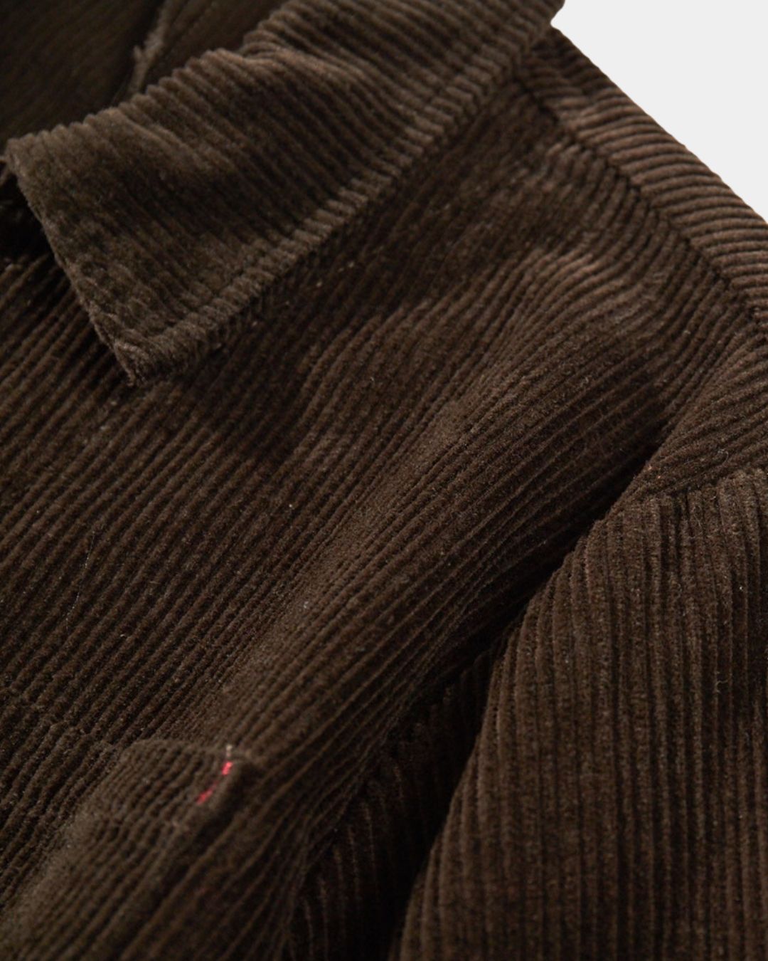 Close-up of a brown ribbed cotton corduroy chore jacket with a point collar and subtle red bar-tack stitching.
