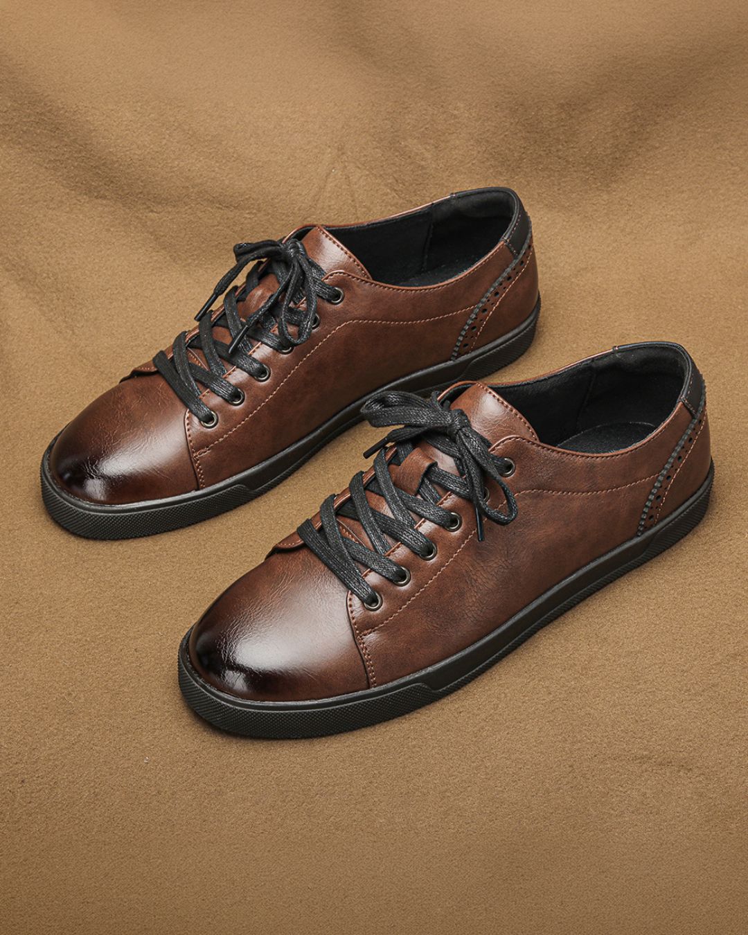 Men’s brown low-top dress sneakers with black laces, burnished finish, and brogue heel detailing on a rubber sole.