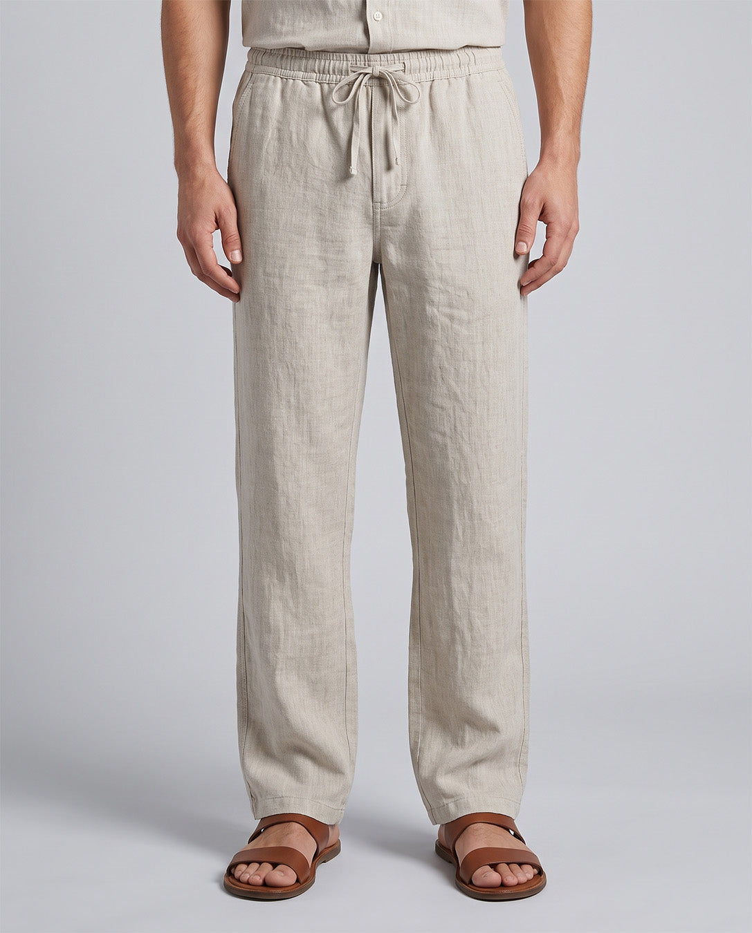 A person wearing light beige drawstring linen pants and brown leather slide sandals, shown from the waist down against a light gray studio background.