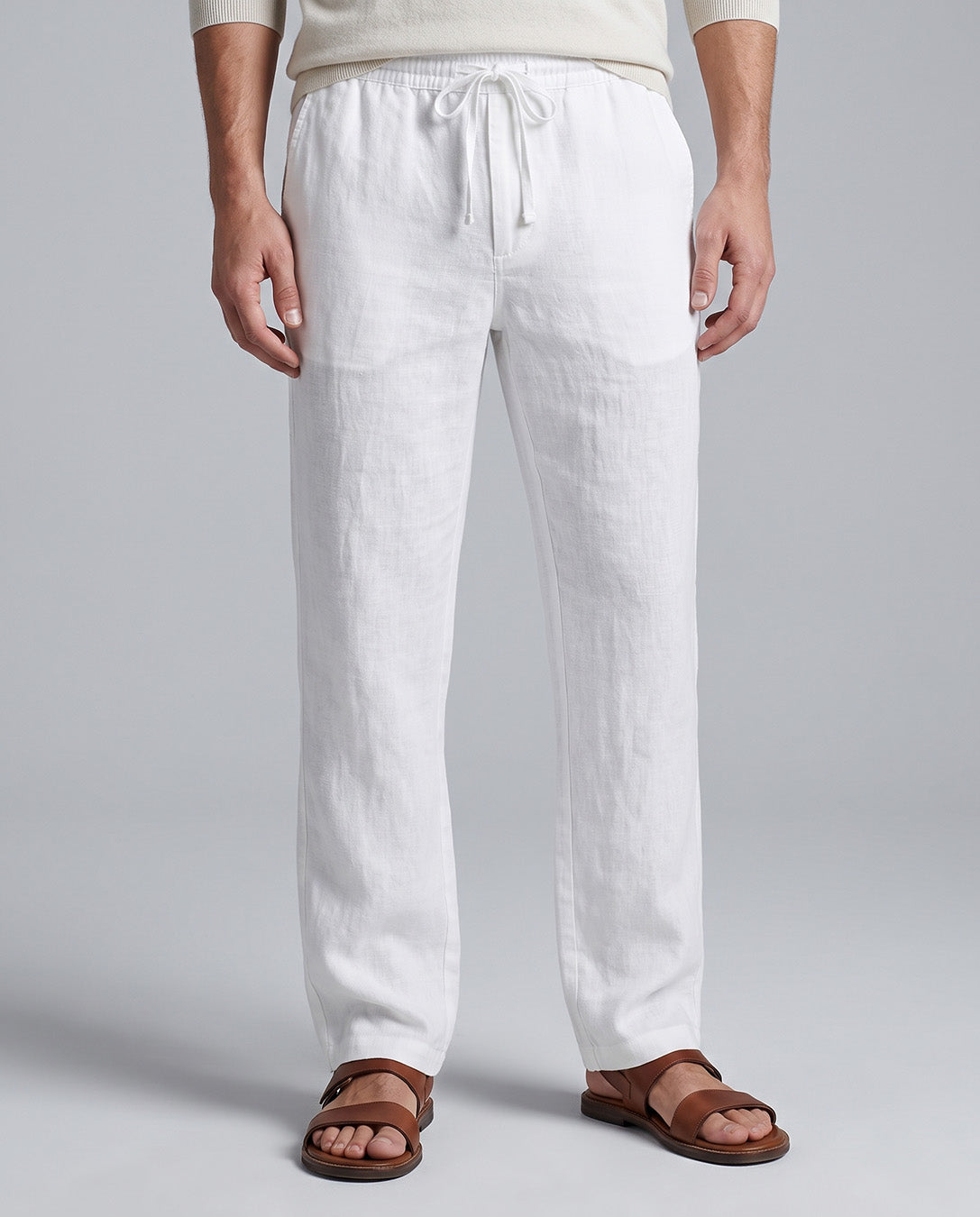 White linen drawstring pants worn by a model, paired with brown leather slide sandals, shown from the waist down on a light gray studio background.