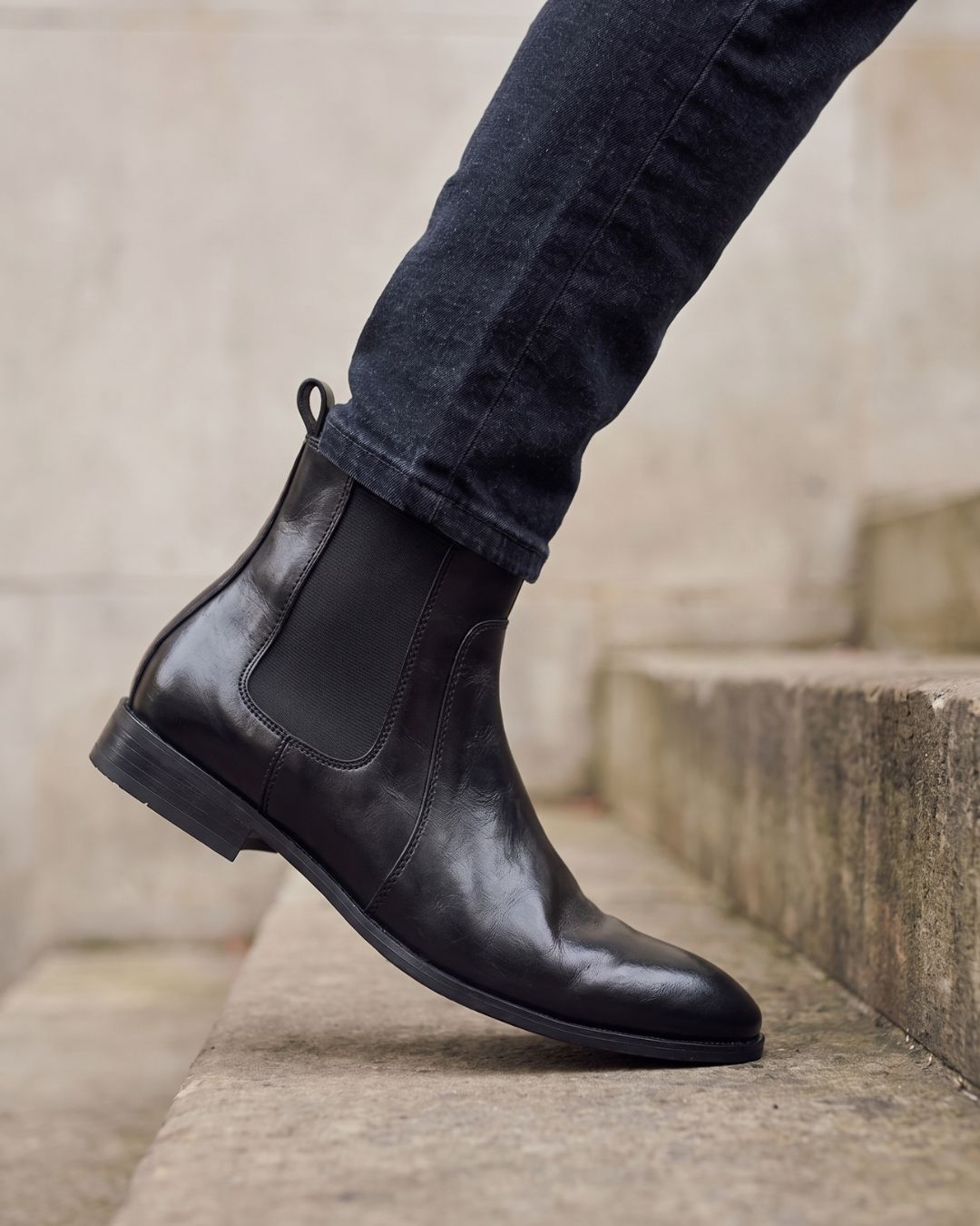 A black leather Chelsea boot worn with dark cuffed jeans, shown on a person stepping onto stone steps.