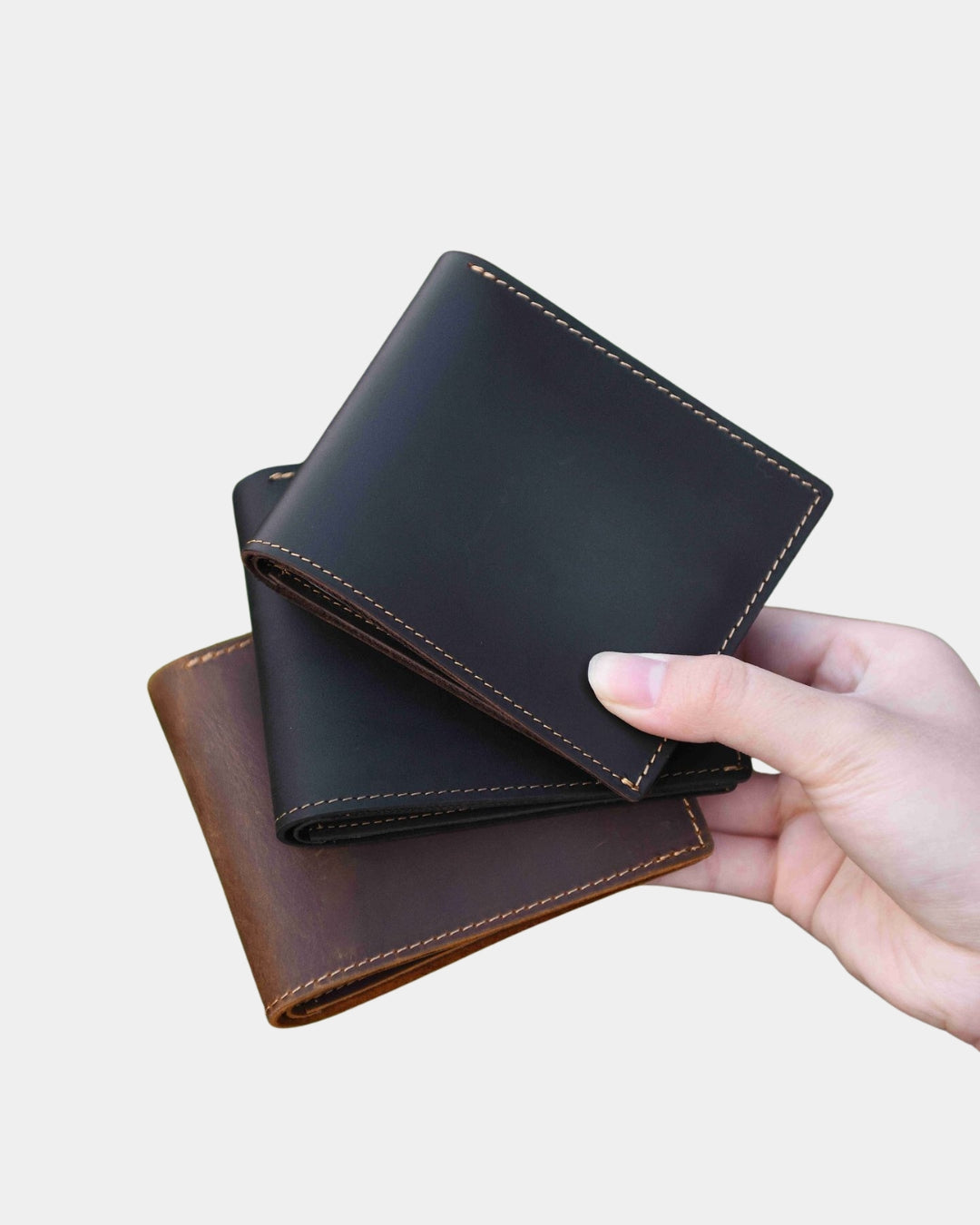 Hand holding two bifold wallets in brown and black Crazy Horse leather with contrast stitching.