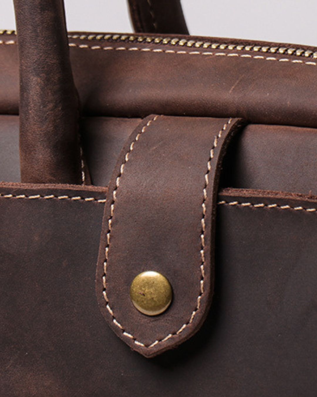 Close-up of a dark brown leather bag featuring a strap closure with a brass snap button and light-colored contrast stitching.