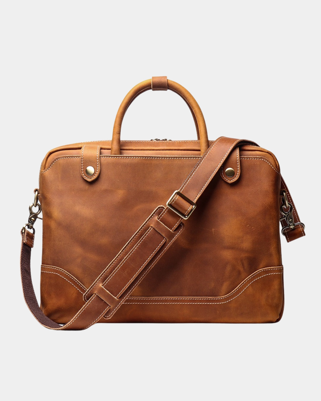 A cognac brown distressed leather briefcase with a top handle, adjustable shoulder strap, and brass hardware, shown against a light gray background.