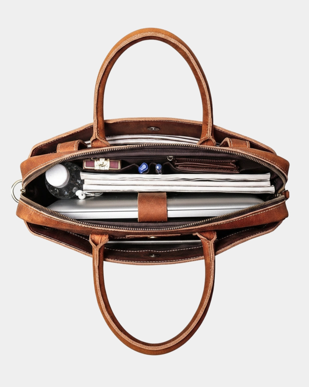 A cognac brown leather tote bag shown open from a top-down view, filled with a laptop, notebook, and small accessories, on a light gray background.