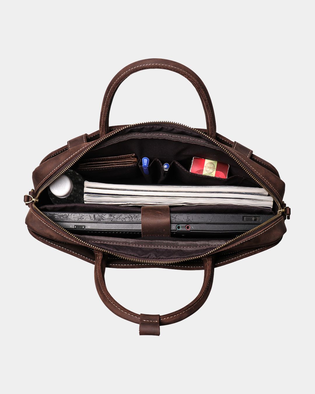 An open brown leather briefcase or satchel shown from above, revealing its interior compartments filled with a laptop, notebook, pens, and other accessories, on a light gray background.