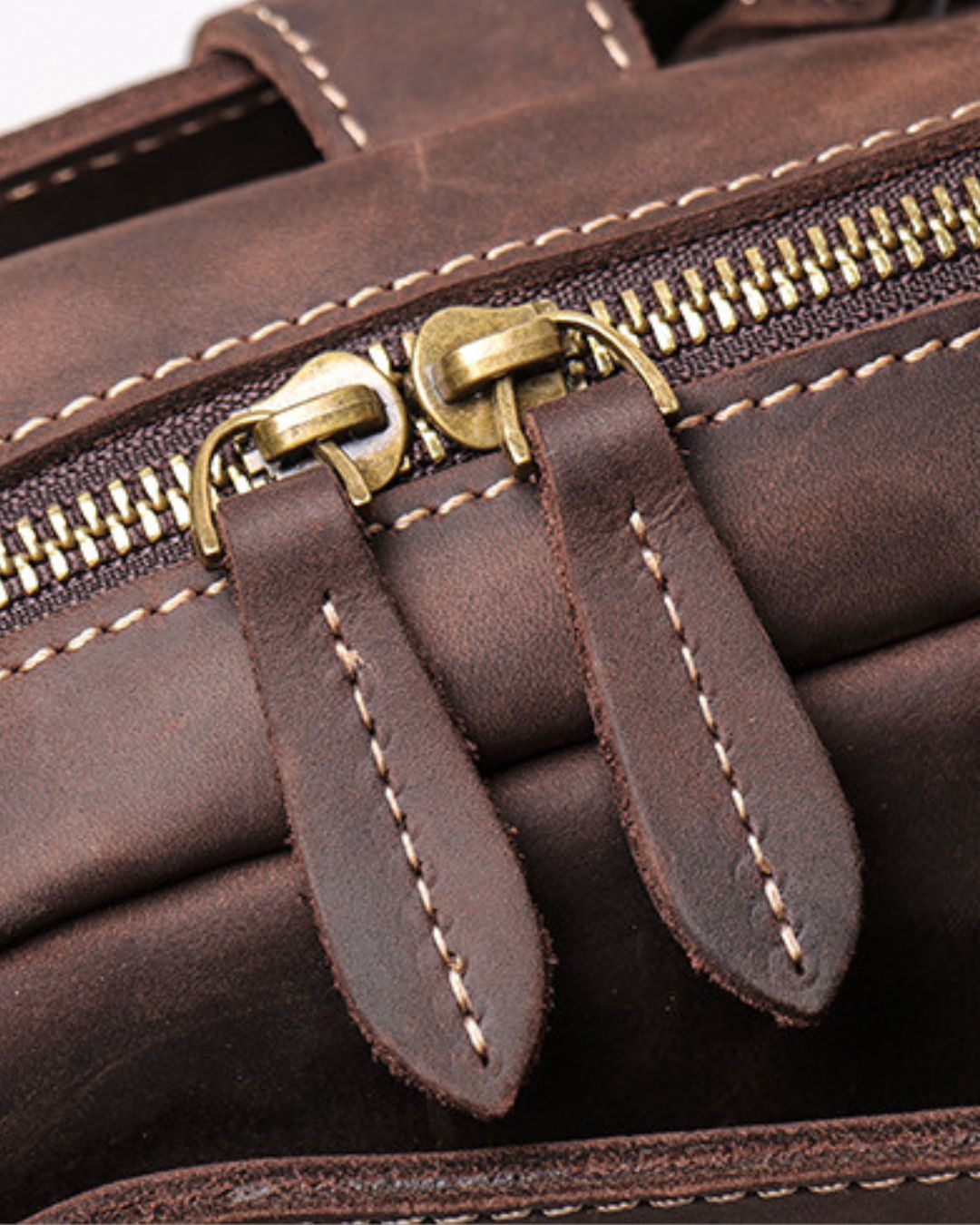 Close-up of a dark brown leather bag with brass zipper pulls and white contrast stitching.