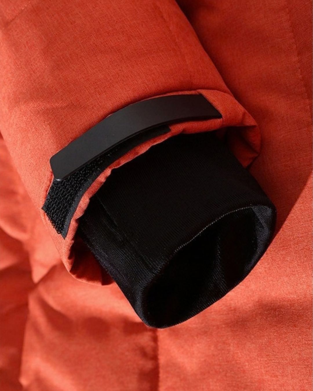 Close-up of a rust-orange puffer ski jacket sleeve featuring an adjustable velcro strap and a black inner stretch cuff.