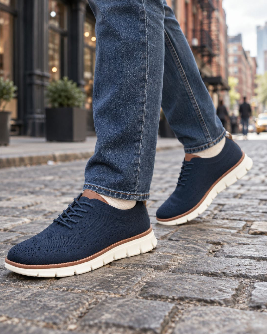 Navy blue mesh dress sneakers with brown leather accents and white EVA soles worn with jeans on a city street.