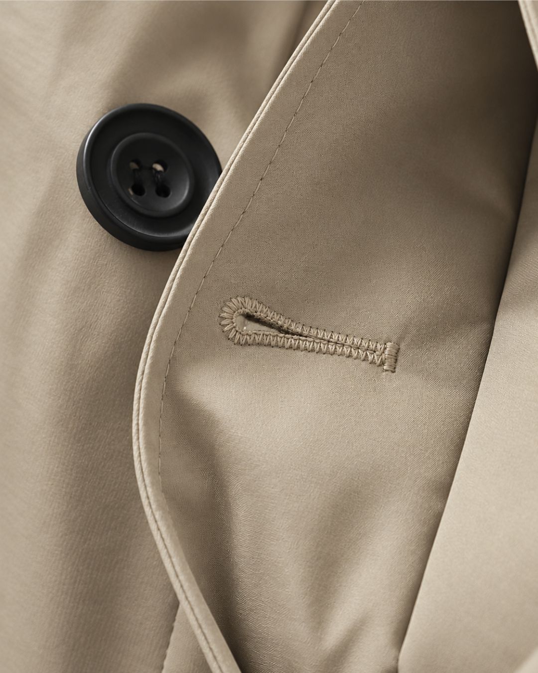 Close-up of a beige men's trench coat featuring a smooth fabric texture, a black button, and detailed stitching.