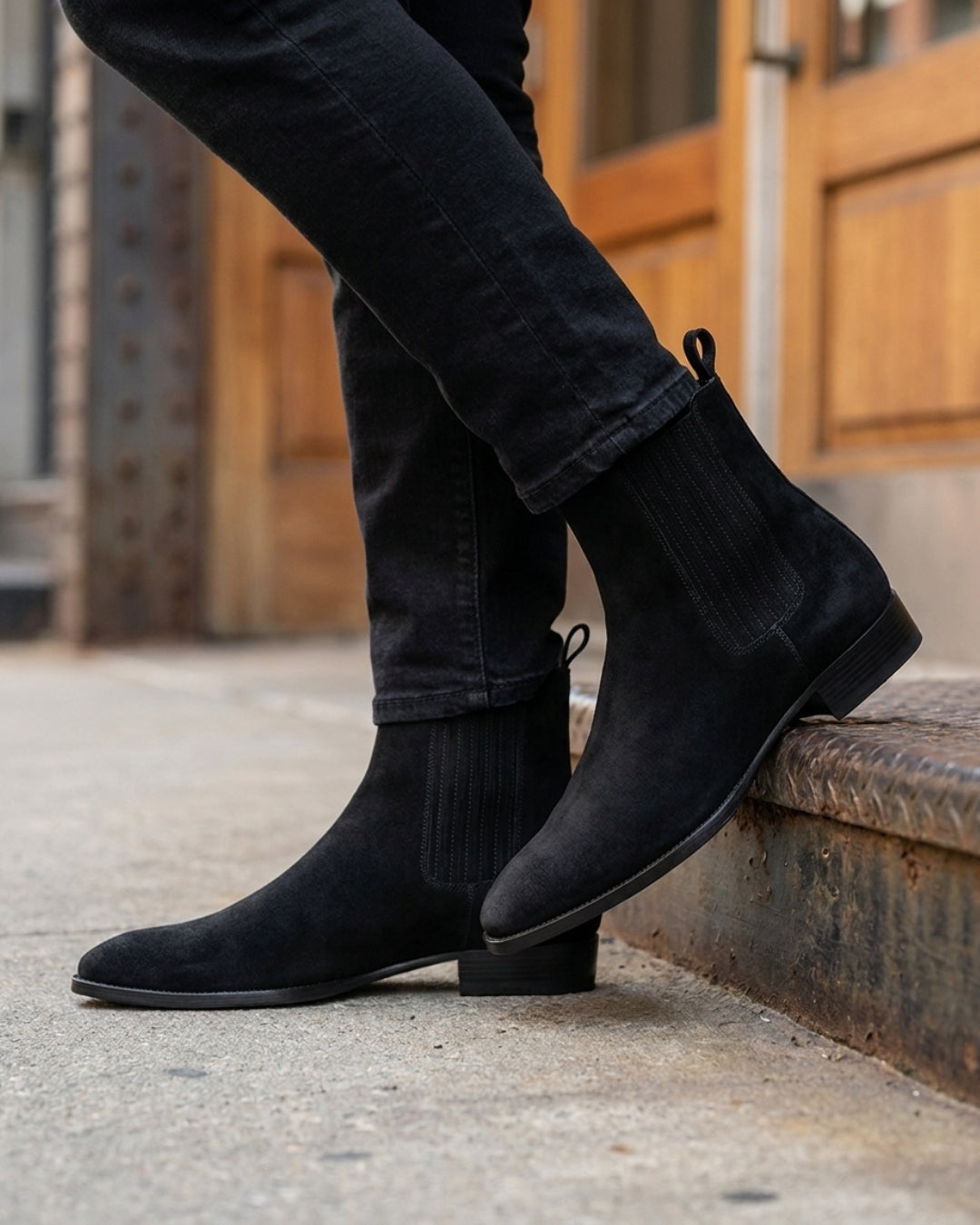 Men’s black suede Chelsea boots with a pointed toe, elastic side panels, and a low heel worn with black jeans.