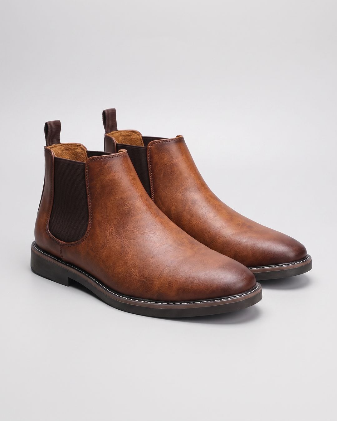A pair of men's brown leather Chelsea boots with dark brown elastic side panels, shown on a light gray studio background.