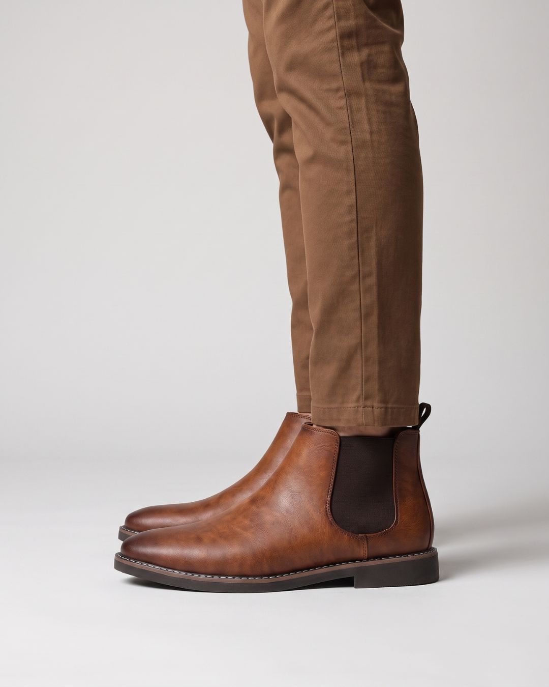 A pair of men's brown leather Chelsea boots worn with brown trousers, shown from a side profile against a light gray studio background.