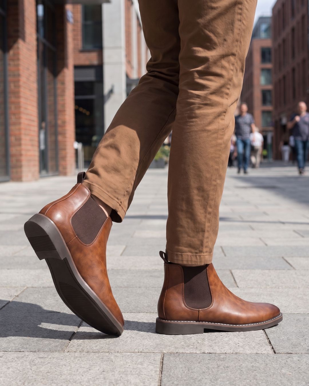 A person wearing tan pants and brown leather Chelsea boots with dark elastic side panels, standing on a paved city walkway with buildings in the background.