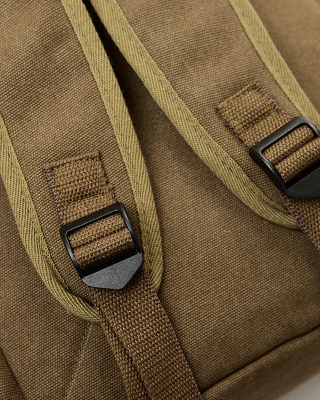 Close-up of a khaki canvas backpack showing the shoulder straps, webbing, and black plastic buckle details.